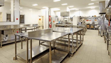 Large commercial kitchen with stainless steel tables and appliances, bright overhead lighting.